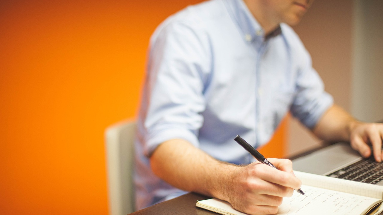 Homme prenant des notes dans un cahier devant un ordinateur portable.