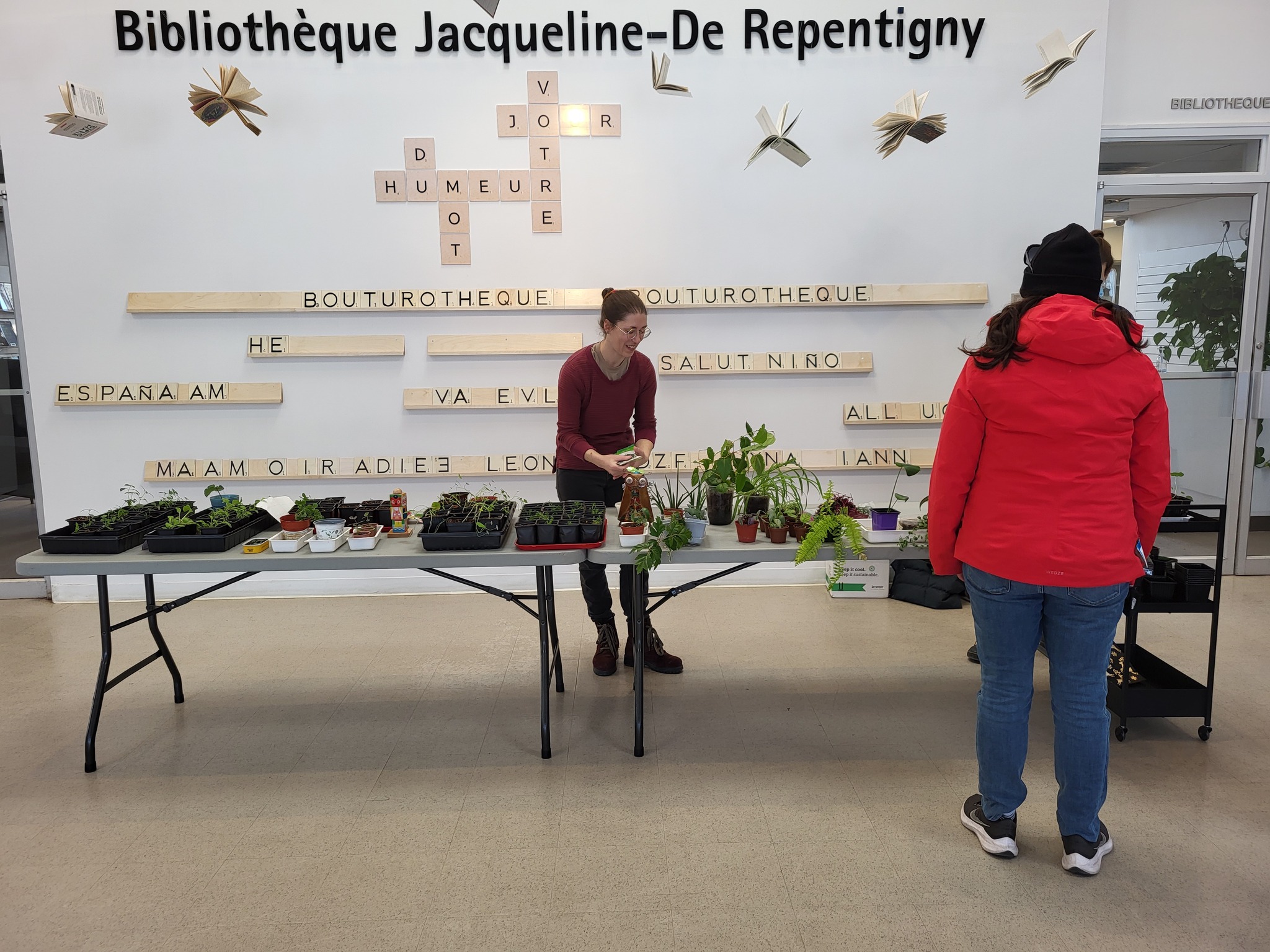 Un femme place des accessoires sur une table sur laquelle se trouvent de nombreuses boutures. Une autre femme se dirige vers la table.