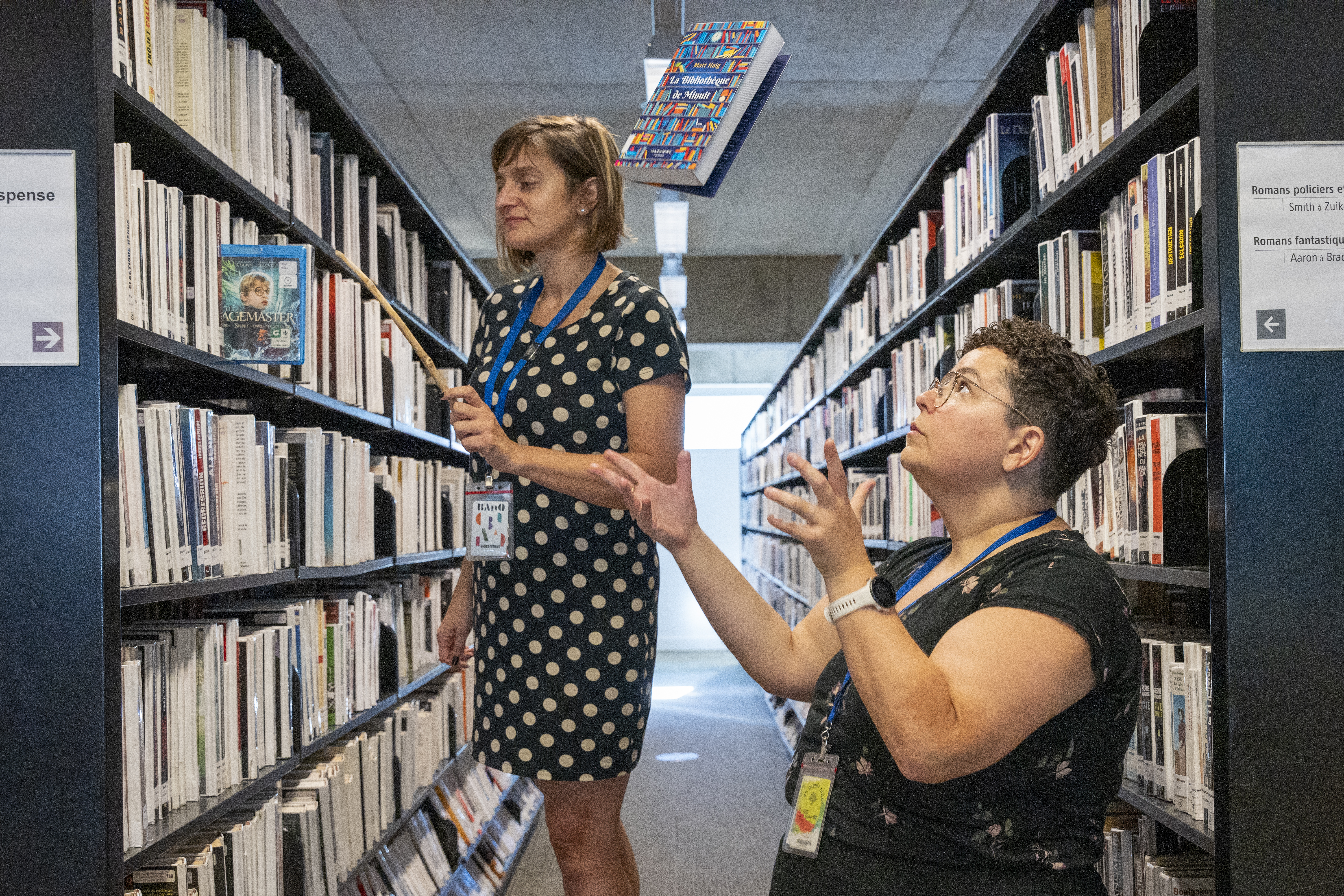 Deux femmes dans une bibliothèque. Un boîtier de DVD et un livre semblent voler.
