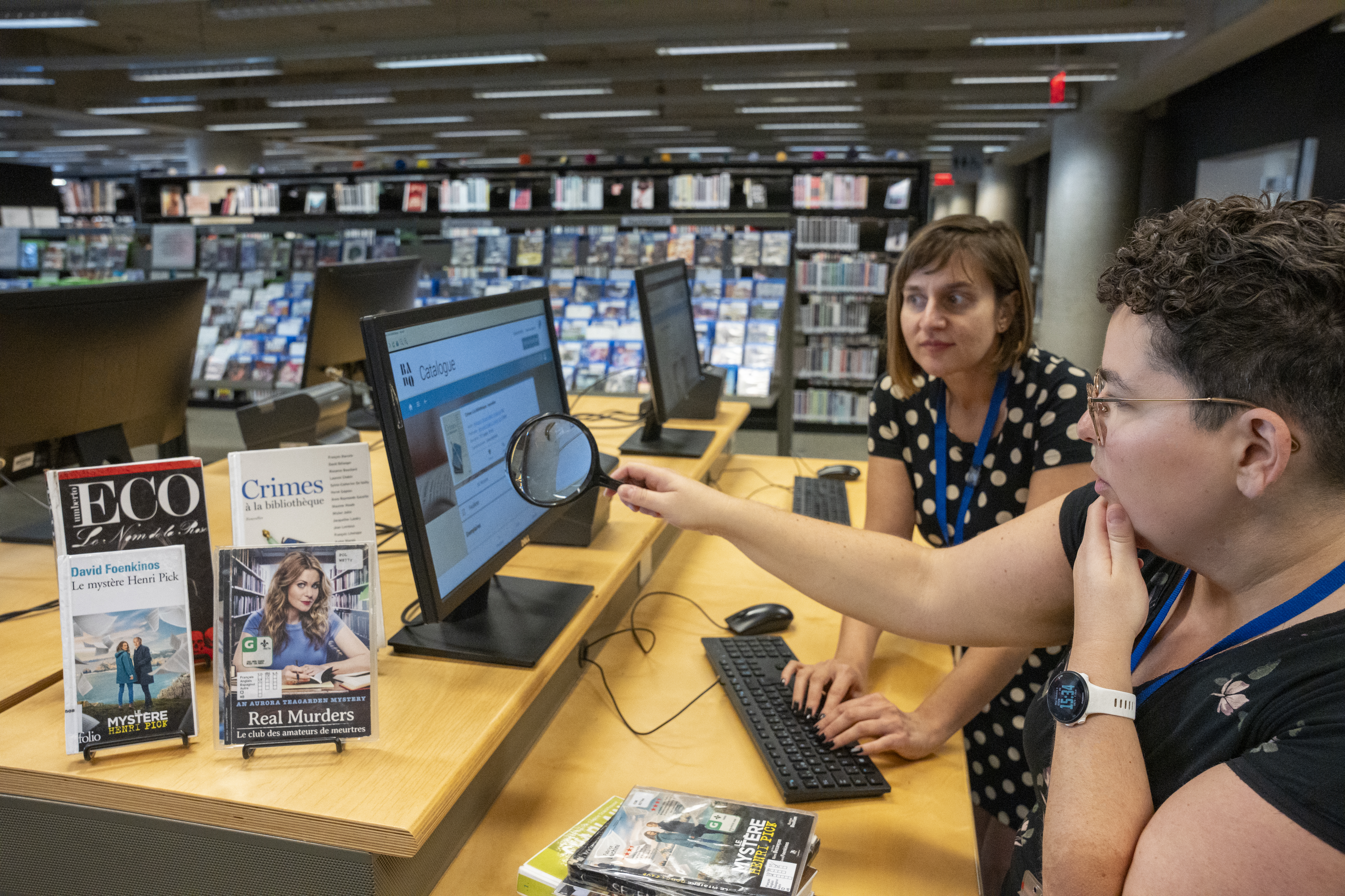 Deux femmes devant un ordinateur. L'une d'elles tient une loupe devant l'écran. À côté, trois livres : « Le nom de la rose », « Crimes à la bibliothèque », « Le mystère Henri Pick » et le film « Aurora Teagarden, Real Murders ».