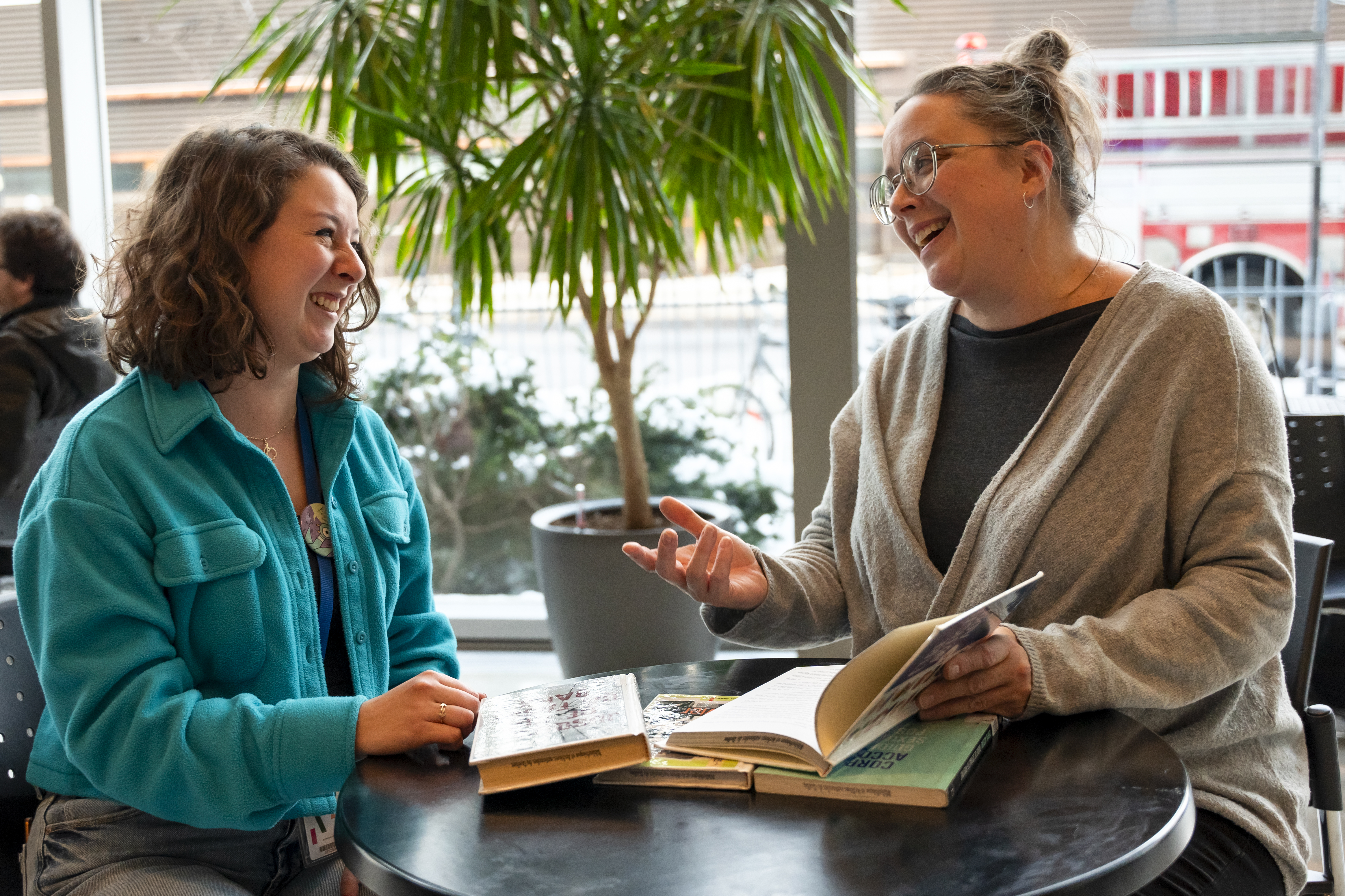 Deux femmes assises à un table où sont déposés des livres discutent.