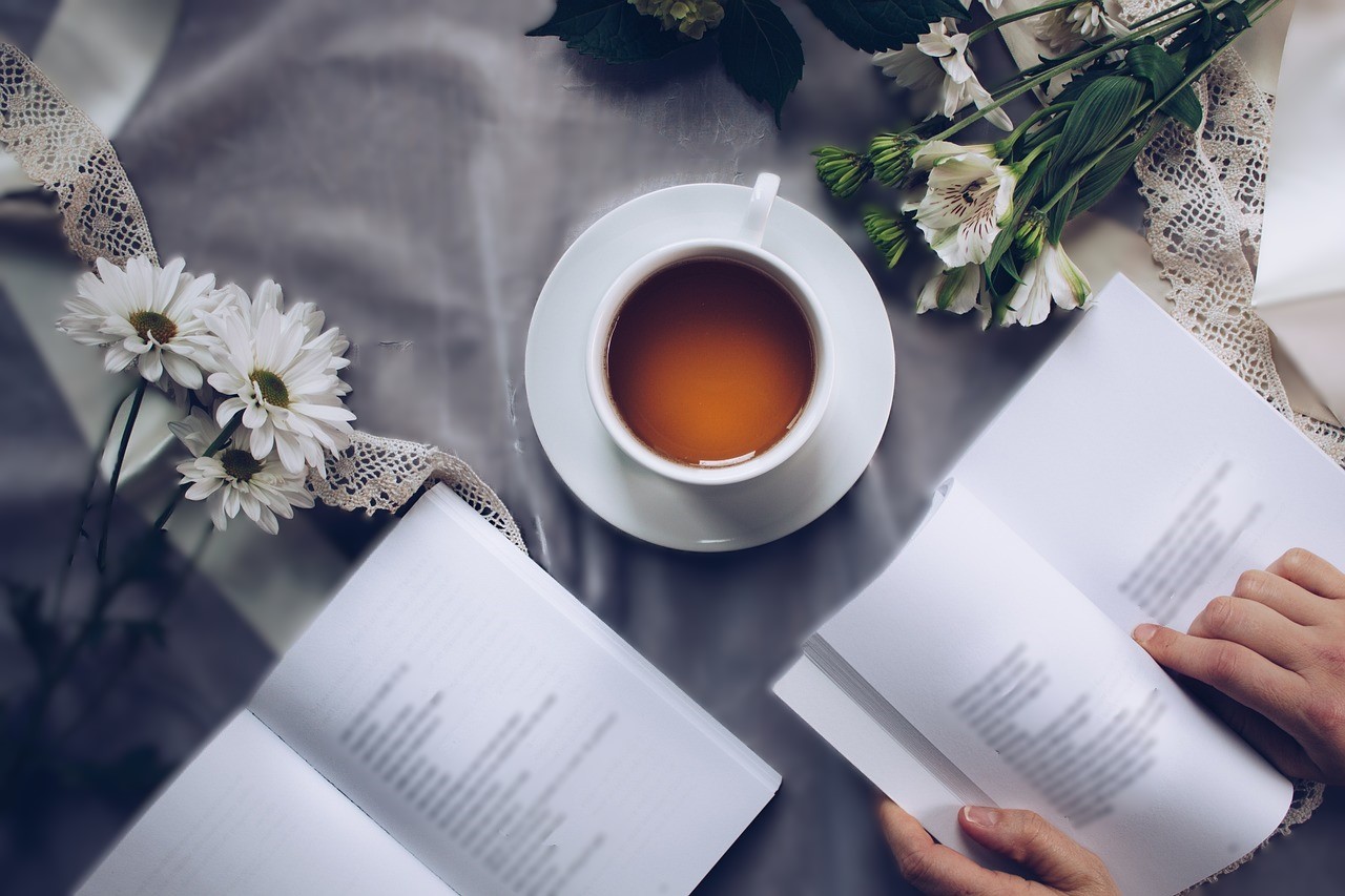 Une personne dont on ne voit que les mains déposées sur les pages est en train de lire un livre qui repose près d'un autre livre ouvert, d'une tasse de thé et de marguerites
