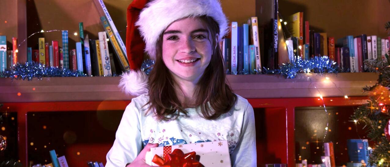 Petite fille avec un chapeau de Noël qui sourit en ouvrant une boîte cadeau entourée de livres et de cadeaux au pied du sapin.