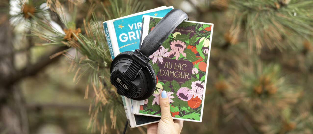 Une main tient deux livres et un casque d'écoute devant un conifère.