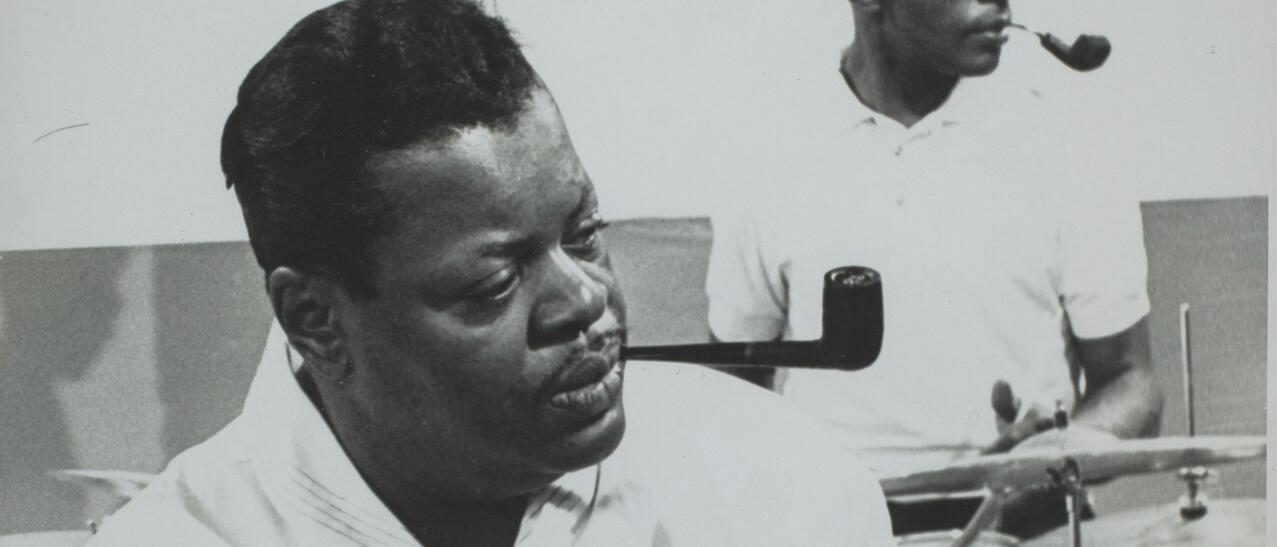 Oscar Peterson, pipe au bec, jouant du piano. Un batteur est visible en arrière-plan. Il porte des lunettes fumées.