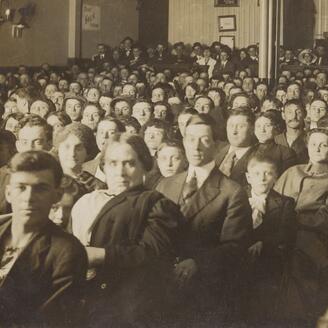 Des dizaines de personnes assises dans une salle accueillant des spectacles et des projections. Les spectateurs fixent l’objectif. 