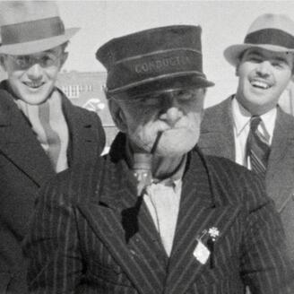 Trois personnes en manteaux et chapeaux posent à l’extérieur devant un bâtiment en arrière-plan.